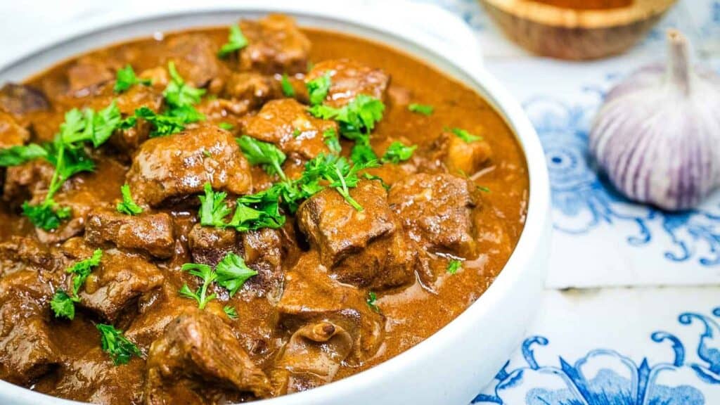 A bowl of beef stew garnished with fresh parsley sits on a blue and white patterned surface near a clove of garlic.