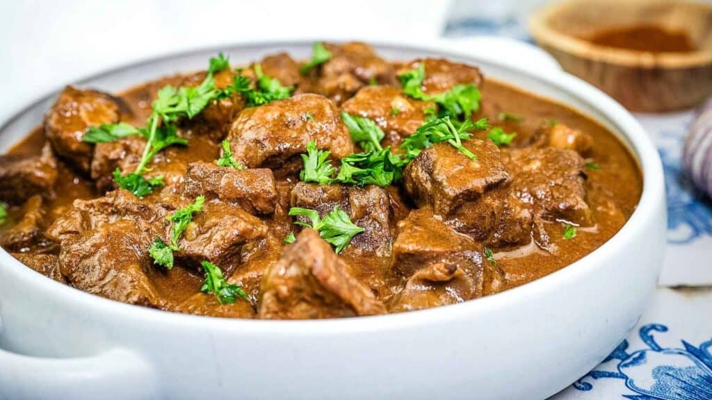 A white bowl filled with beef stew in brown gravy, garnished with fresh parsley.