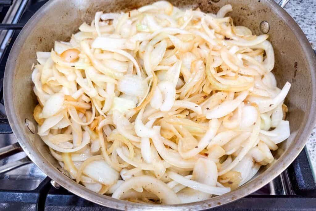 Sliced onions cooking and beginning to brown in a large metal pan on a stovetop.