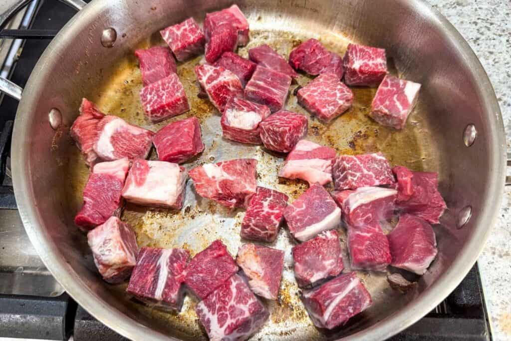 Cubes of raw beef are browning in a stainless steel pan on a stove, with visible marbling and some pieces starting to sear.