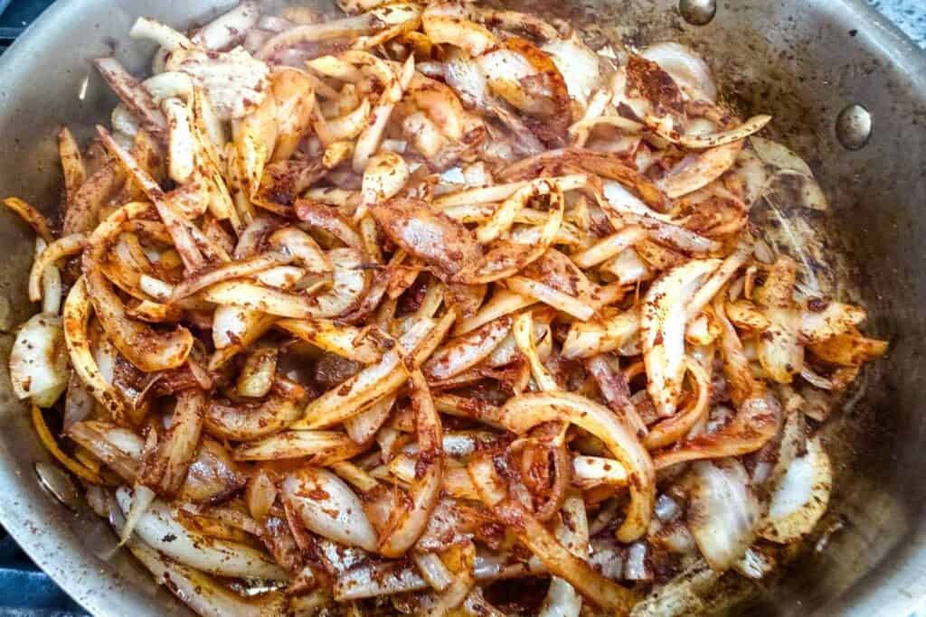 Sliced onions being sautéed in a pan with spices, showing a mix of golden brown and translucent pieces.