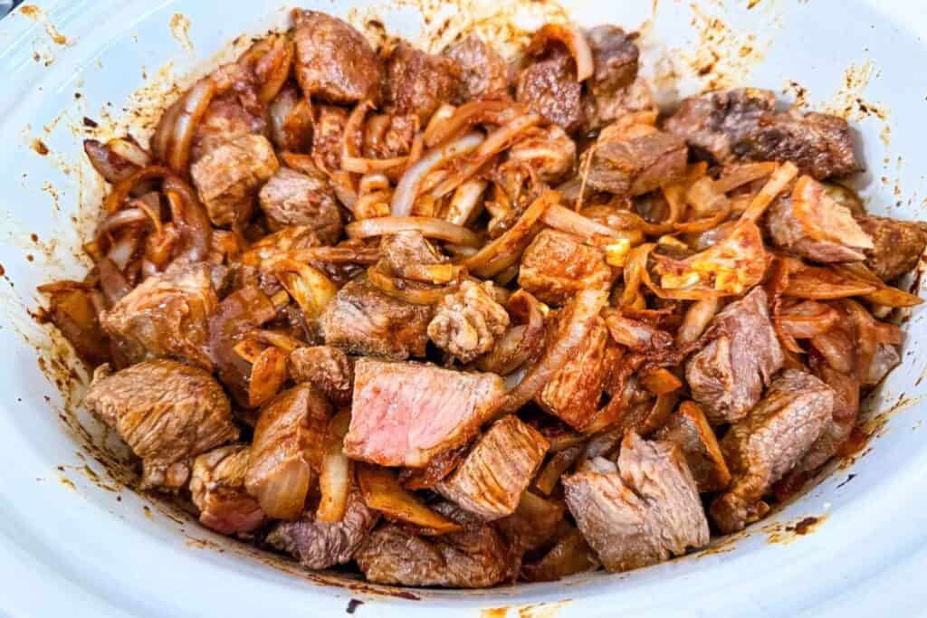 Chunks of cooked beef and sliced onions mixed with spices in a white slow cooker.