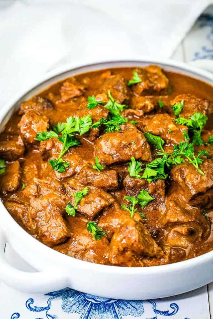 A white bowl filled with beef stew in a thick brown sauce, garnished with chopped fresh parsley.