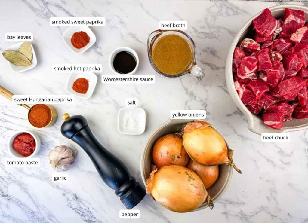 Ingredients for beef stew on a marble surface: beef chuck, onions, beef broth, bay leaves, tomato paste, garlic, pepper, paprika, Worcestershire sauce, salt, and smoked paprikas.