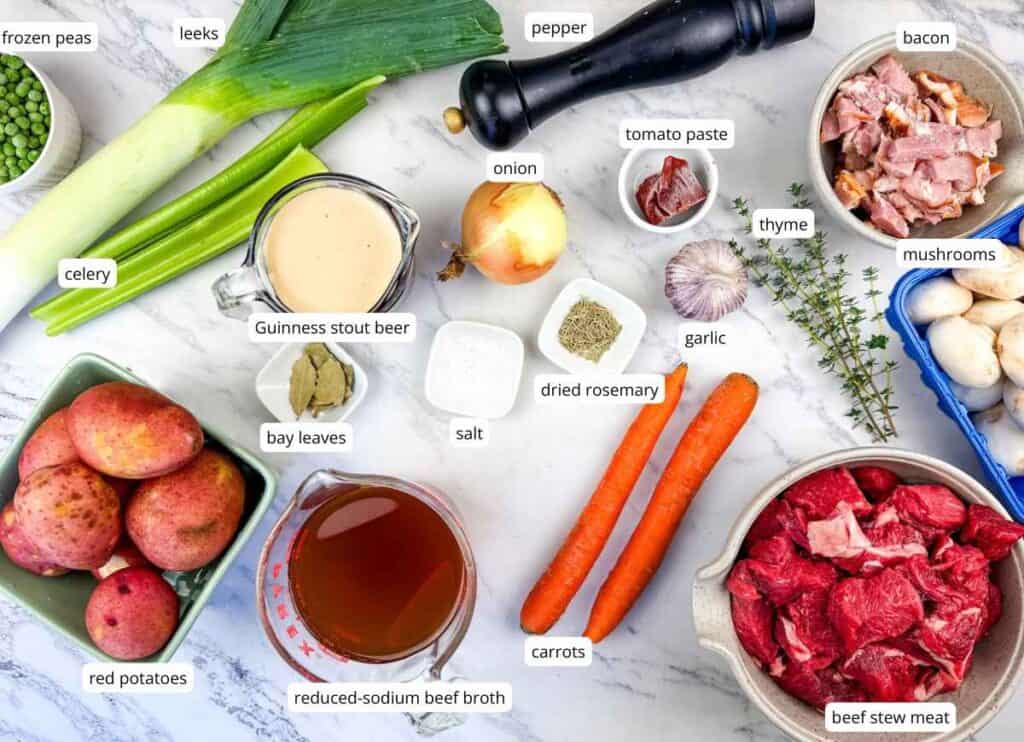 Ingredients for a beef stew recipe arranged on a counter, including red potatoes, leeks, celery, Guinness beer, beef broth, carrots, bacon, mushrooms, beef, and various seasonings, all labeled.