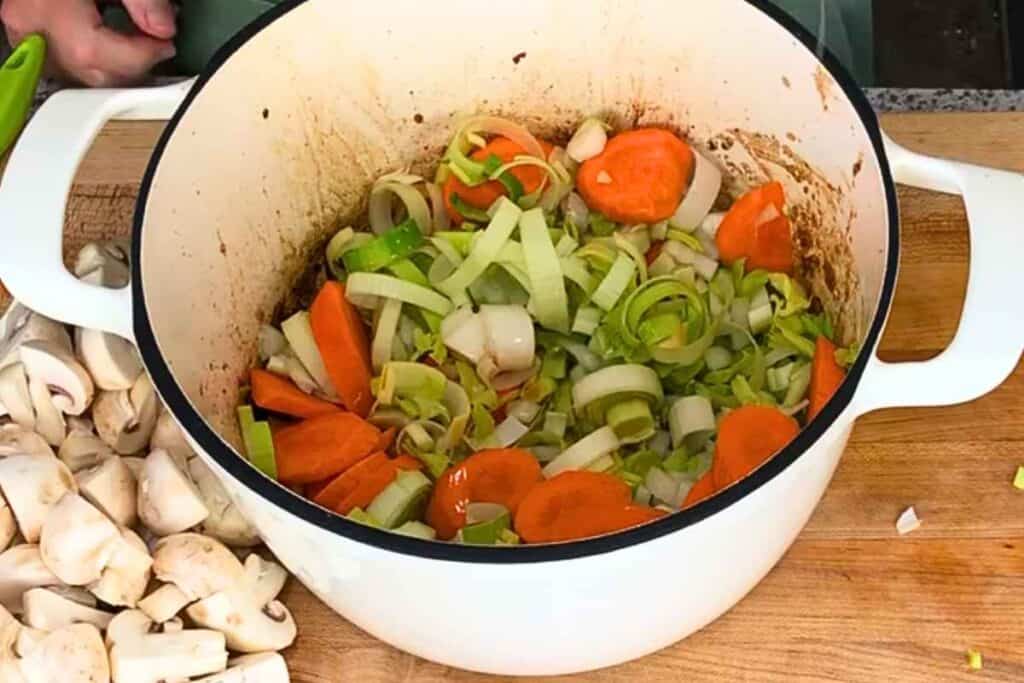 Sliced carrots and leeks sautéing in a white enameled cast iron pot next to a pile of chopped mushrooms.