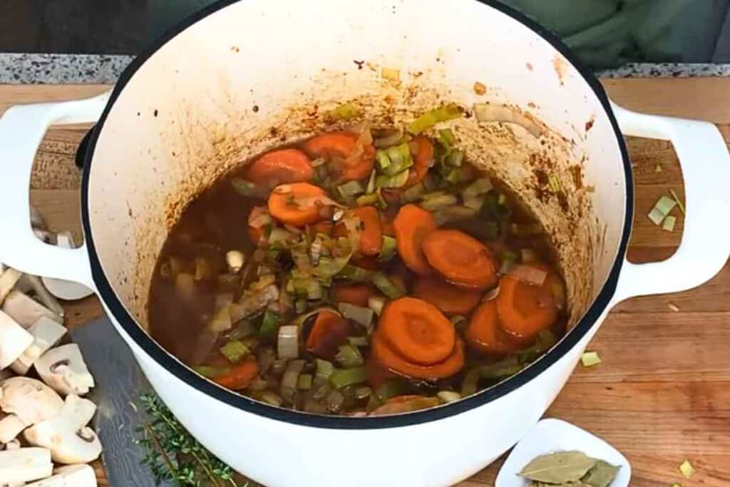 A white Dutch oven containing a simmering broth with sliced carrots, celery, and onions, with a pile of sliced mushrooms and herbs on the counter nearby.