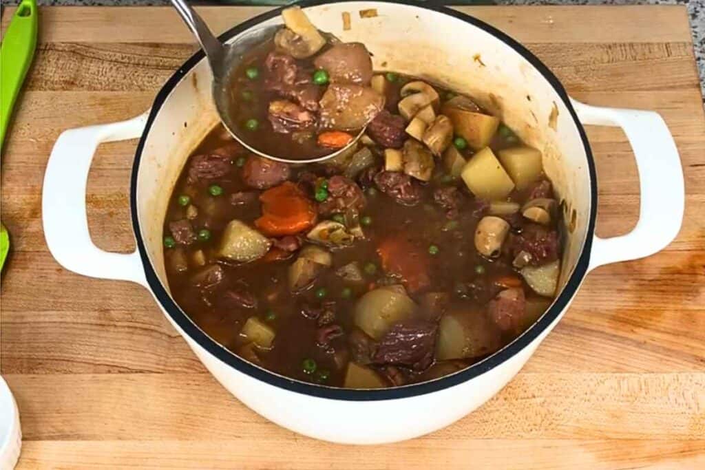 A ladle lifts beef stew with potatoes, carrots, peas, and mushrooms from a large white Dutch oven on a wooden cutting board.