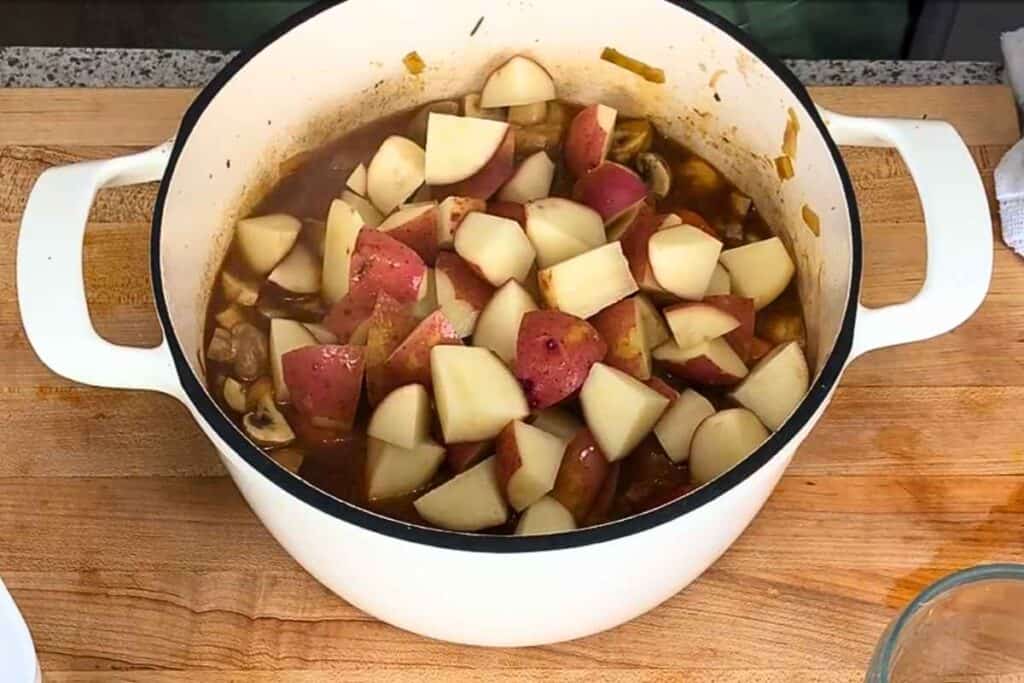 A white enameled pot on a wooden cutting board filled with stew containing chunks of red-skinned potatoes and mushrooms.