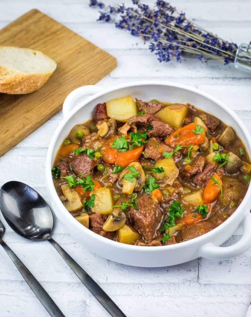 A white bowl filled with beef stew containing chunks of meat, potatoes, carrots, mushrooms, and peas, garnished with parsley.