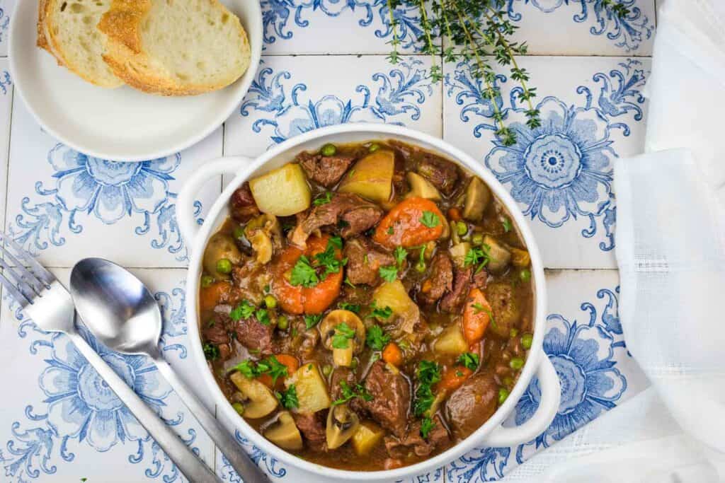Beef stew with potatoes, carrots, mushrooms, and peas in a white bowl, served with sliced bread.